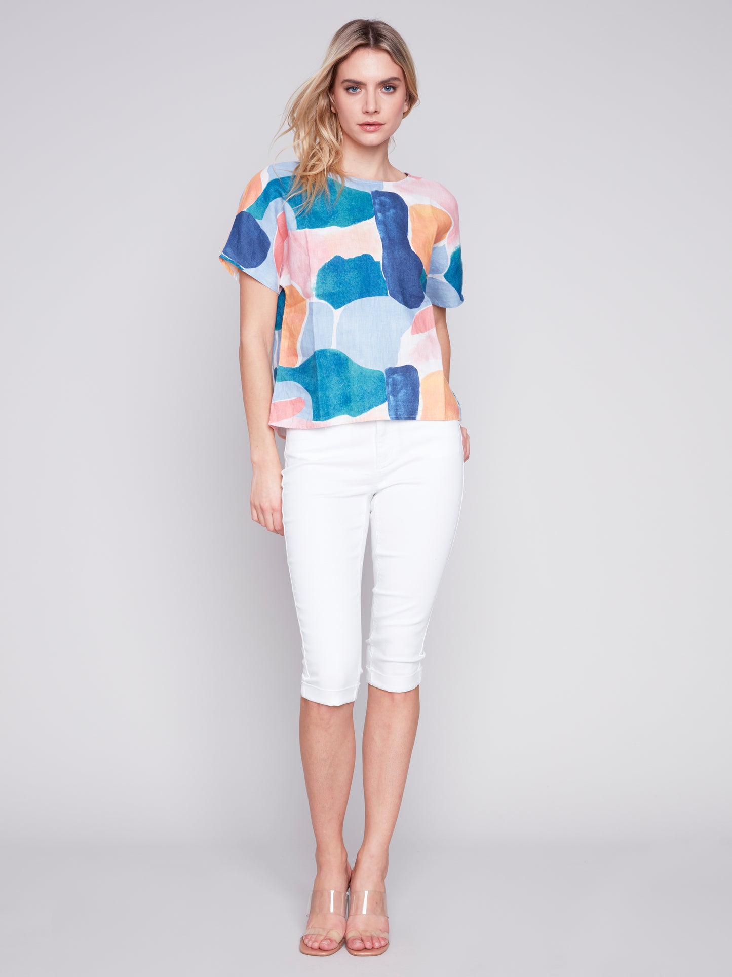 A blonde woman in a colorful "Charlie B Dolman Linen Printed Top" and white pants, standing against a light gray background.