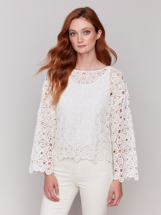 A person with long hair, draped in a Charlie B Cotton Blend Crochet Top and white pants, stands gracefully against a neutral backdrop.