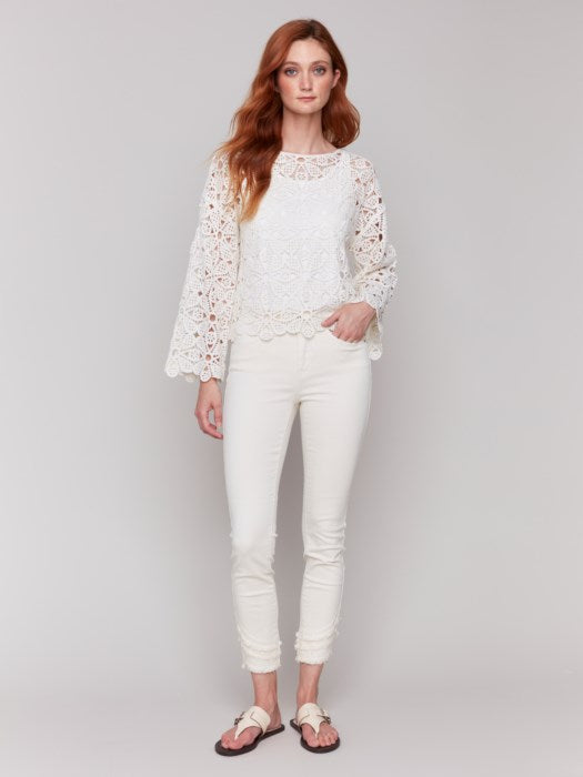 A person with long hair, draped in a Charlie B Cotton Blend Crochet Top and white pants, stands gracefully against a neutral backdrop.