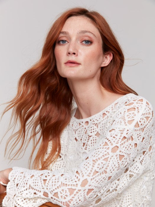 A person with long hair, draped in a Charlie B Cotton Blend Crochet Top and white pants, stands gracefully against a neutral backdrop.