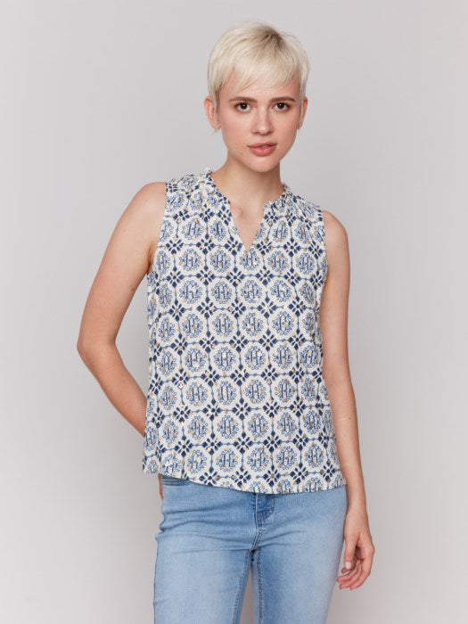 A person with short blonde hair wears a Charlie B Printed Sleeveless Ruffle Neck Top, light blue capri jeans, and beige sandals, standing against a plain backdrop.