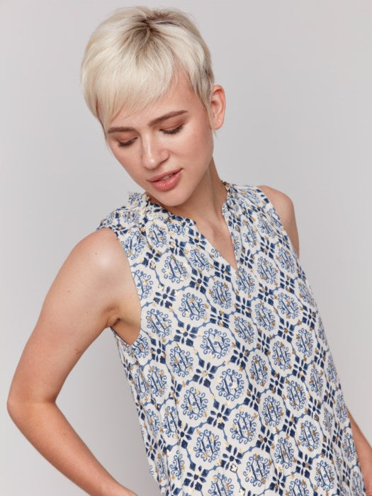 A person with short blonde hair wears a Charlie B Printed Sleeveless Ruffle Neck Top, light blue capri jeans, and beige sandals, standing against a plain backdrop.