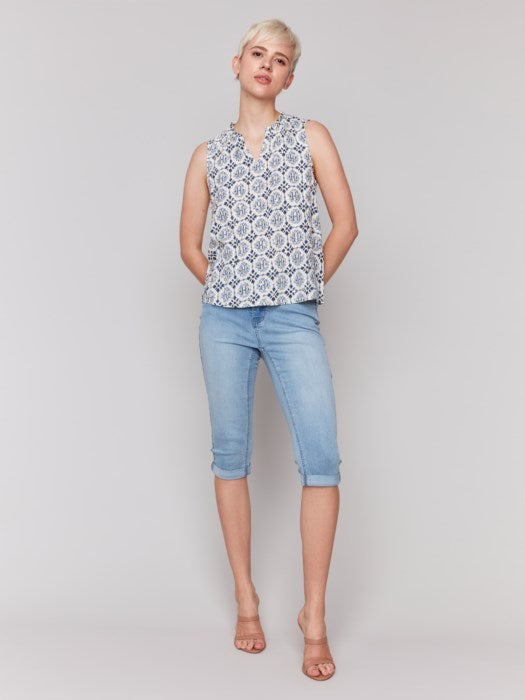 A person with short blonde hair wears a Charlie B Printed Sleeveless Ruffle Neck Top, light blue capri jeans, and beige sandals, standing against a plain backdrop.