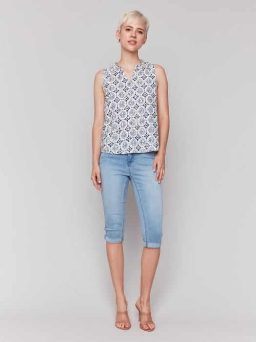 A person with short blonde hair wears a Charlie B Printed Sleeveless Ruffle Neck Top, light blue capri jeans, and beige sandals, standing against a plain backdrop.