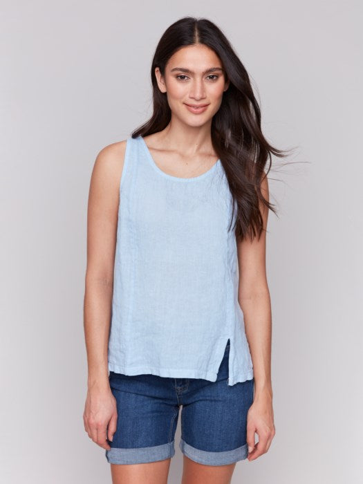 A fashionista with long dark hair models the Charlie B Sleeveless Linen Top with Slit in light blue paired with denim shorts, standing confidently against a plain gray background.