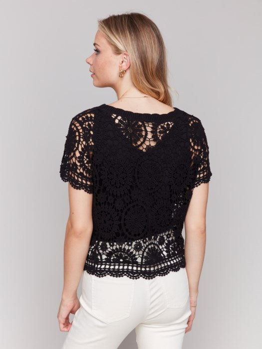 A person poses fashion-forward in a Charlie B Short Sleeve Crochet Bolero Top, paired with white pants, against a plain background.