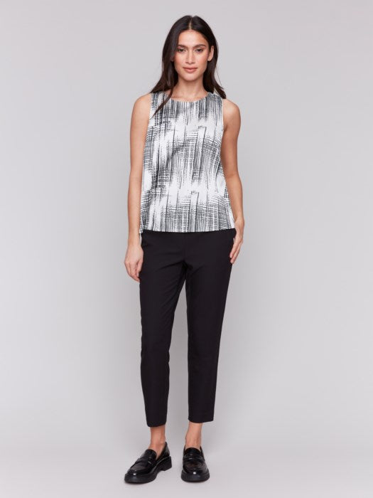 A woman with long hair models the Charlie B Printed Satin Sleeveless Top—stylishly bra-friendly and black-and-white printed—paired with black pants, set against a plain gray background.