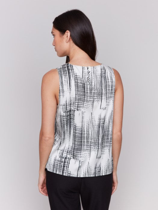 A woman with long hair models the Charlie B Printed Satin Sleeveless Top—stylishly bra-friendly and black-and-white printed—paired with black pants, set against a plain gray background.