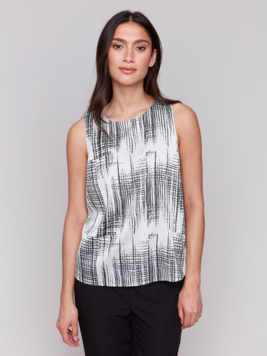 A woman with long hair models the Charlie B Printed Satin Sleeveless Top—stylishly bra-friendly and black-and-white printed—paired with black pants, set against a plain gray background.