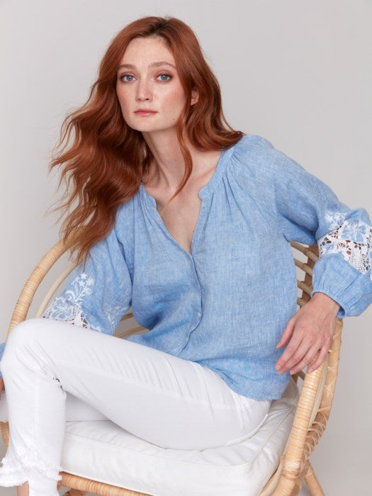 A person with long red hair sits in a wicker chair, wearing a Charlie B Button-Front Blouse with Embroidery and white pants, gazing forward.