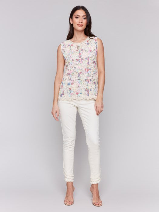 A person is wearing a Sleeveless Crochet Crew Neck Top by Charlie B, featuring a vibrant pattern, paired elegantly with white pants against a plain gray background.