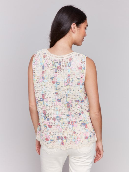 A person is wearing a Sleeveless Crochet Crew Neck Top by Charlie B, featuring a vibrant pattern, paired elegantly with white pants against a plain gray background.