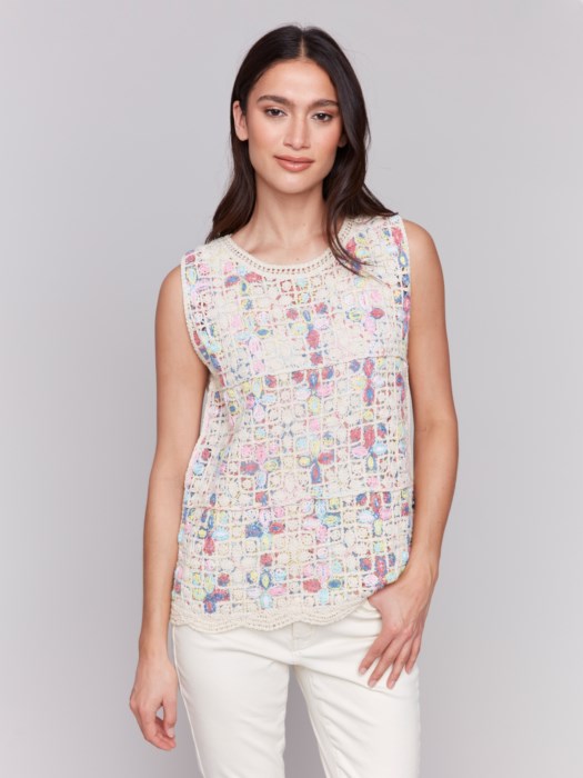 A person is wearing a Sleeveless Crochet Crew Neck Top by Charlie B, featuring a vibrant pattern, paired elegantly with white pants against a plain gray background.