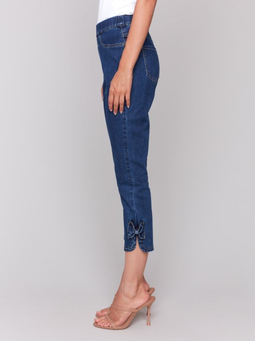 A person exudes effortless style in Charlie B's cropped blue pull-on jeans with bow detail and a white top. They stand against a plain background, hands resting at their sides, feet elegantly adorned in beige heeled sandals.