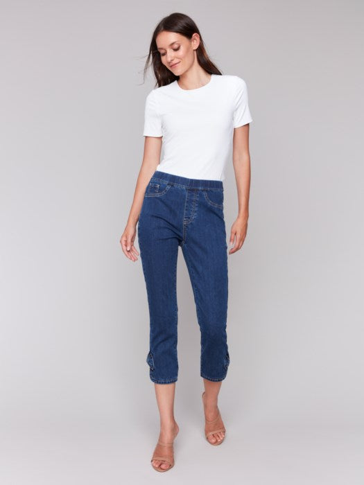 A person exudes effortless style in Charlie B's cropped blue pull-on jeans with bow detail and a white top. They stand against a plain background, hands resting at their sides, feet elegantly adorned in beige heeled sandals.