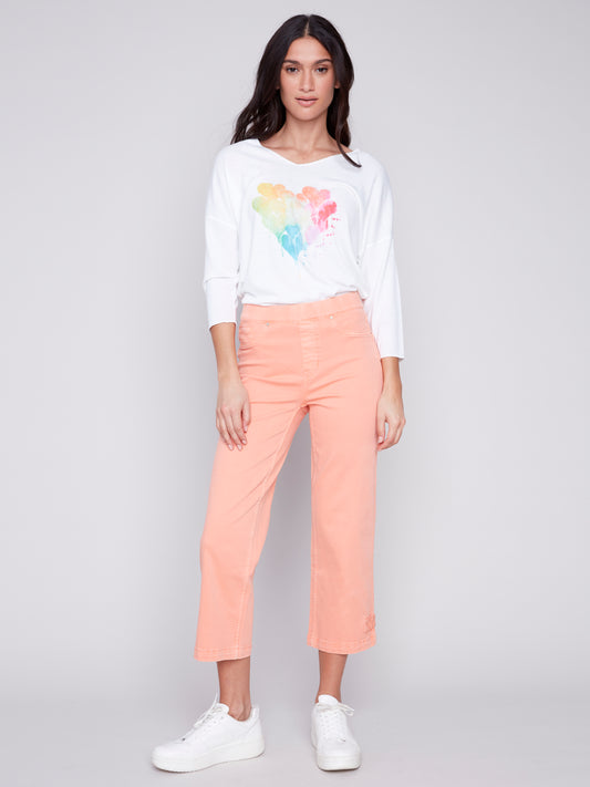 Woman posing in a white t-shirt with a colorful heart design and peach-colored Charlie B Cropped Twill Pants with Hem Tab, paired with white sneakers.