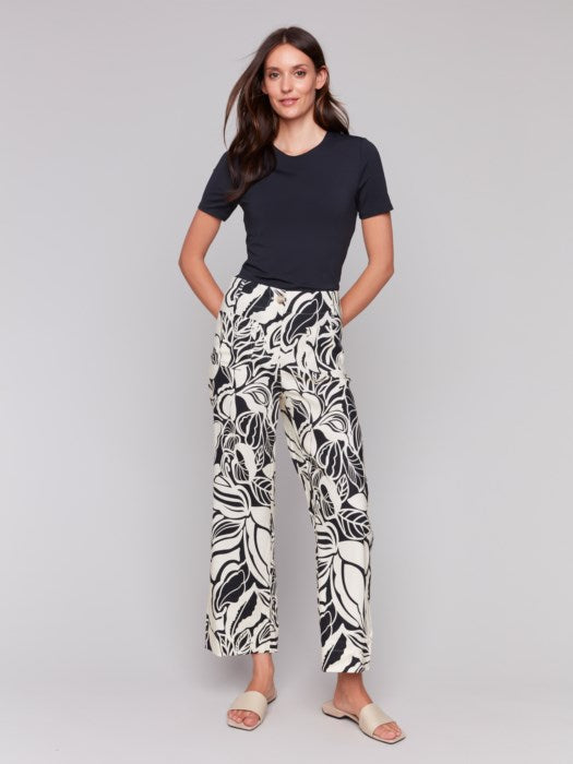 A person is wearing Charlie B's Printed Cropped Linen Blend Pants with a black floral pattern, a black top, and beige open-toe slides while standing against a plain background.