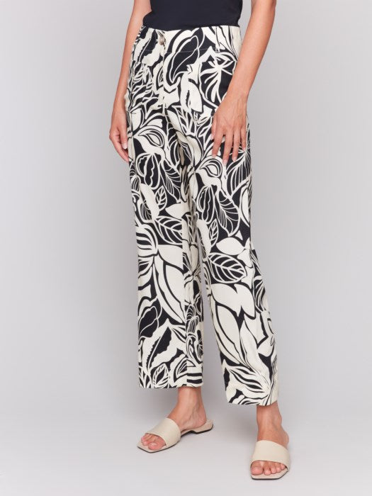 A person is wearing Charlie B's Printed Cropped Linen Blend Pants with a black floral pattern, a black top, and beige open-toe slides while standing against a plain background.