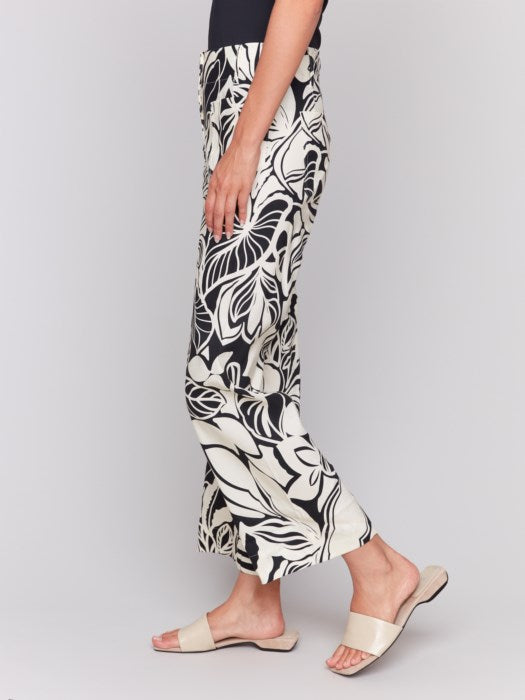 A person is wearing Charlie B's Printed Cropped Linen Blend Pants with a black floral pattern, a black top, and beige open-toe slides while standing against a plain background.