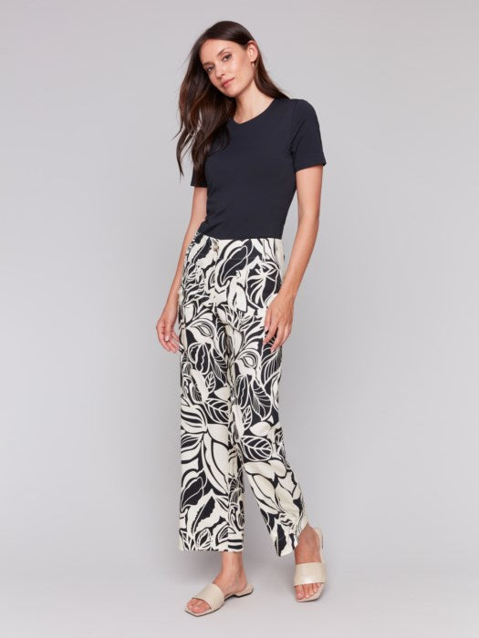 A person is wearing Charlie B's Printed Cropped Linen Blend Pants with a black floral pattern, a black top, and beige open-toe slides while standing against a plain background.