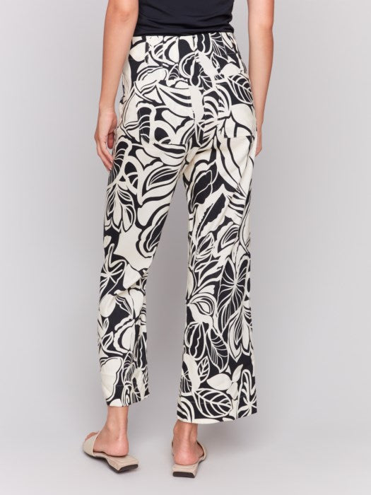 A person is wearing Charlie B's Printed Cropped Linen Blend Pants with a black floral pattern, a black top, and beige open-toe slides while standing against a plain background.