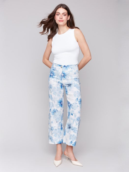 A person is wearing Charlie B's Printed Cropped Linen Blend Pants with a black floral pattern, a black top, and beige open-toe slides while standing against a plain background.