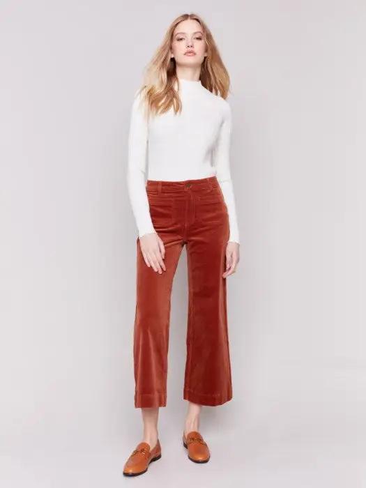 A person wearing a white long-sleeve top, rust-colored Patch Pocket Corduroy Flare Pants from Charlie B made of soft corduroy fabric, and brown loafers stands against a plain background. The high-waisted pants feature a cropped length.