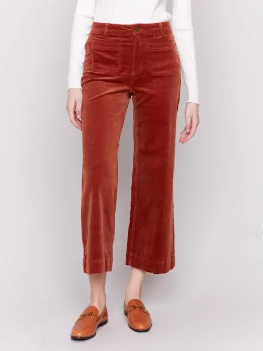 A person wearing a white long-sleeve top, rust-colored Patch Pocket Corduroy Flare Pants from Charlie B made of soft corduroy fabric, and brown loafers stands against a plain background. The high-waisted pants feature a cropped length.