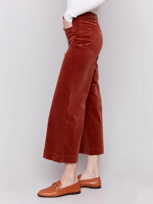 A person wearing a white long-sleeve top, rust-colored Patch Pocket Corduroy Flare Pants from Charlie B made of soft corduroy fabric, and brown loafers stands against a plain background. The high-waisted pants feature a cropped length.