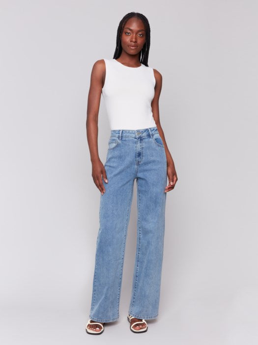 A trendsetter in a white sleeveless top and Charlie B's stylish blue Chloe Wide Leg Jeans stands against a plain gray background.