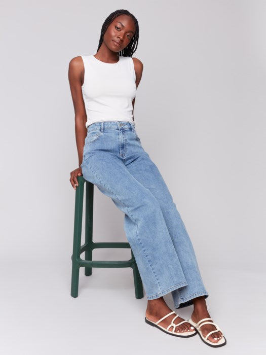 A trendsetter in a white sleeveless top and Charlie B's stylish blue Chloe Wide Leg Jeans stands against a plain gray background.