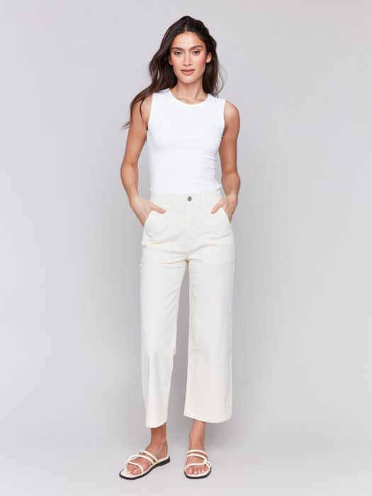 A person in a white sleeveless top and light-colored Cropped Utility Pocket Twill Pants by Charlie B stands against a plain gray background. Their hands rest casually in their pockets, complemented by white sandals.