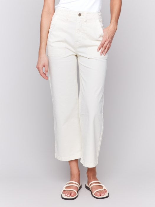 A person in a white sleeveless top and light-colored Cropped Utility Pocket Twill Pants by Charlie B stands against a plain gray background. Their hands rest casually in their pockets, complemented by white sandals.