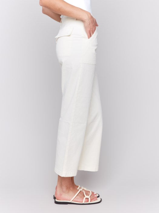 A person in a white sleeveless top and light-colored Cropped Utility Pocket Twill Pants by Charlie B stands against a plain gray background. Their hands rest casually in their pockets, complemented by white sandals.