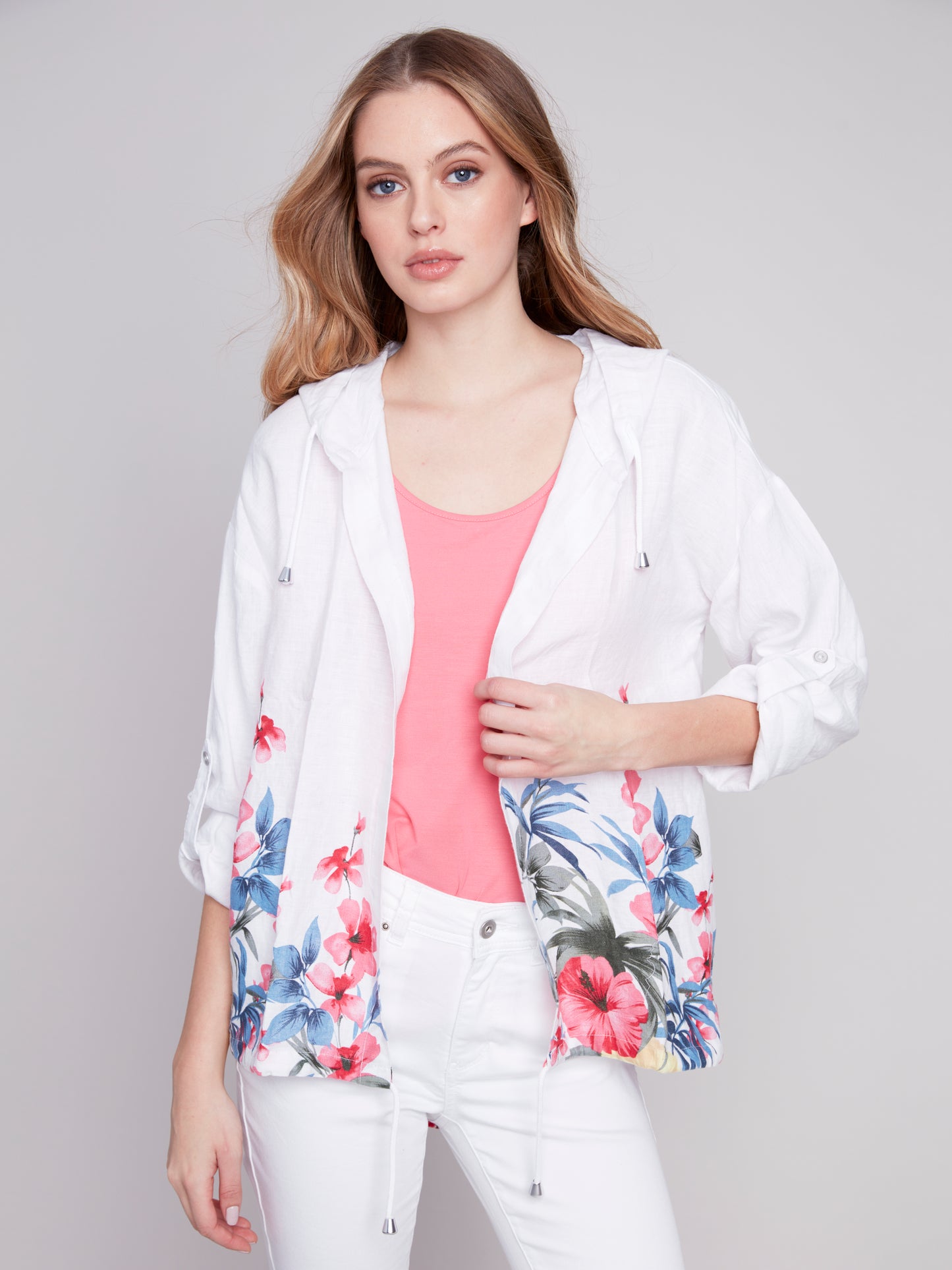 A woman in a white Charlie B floral print linen duster jacket, pink top, and white pants posing against a gray background.