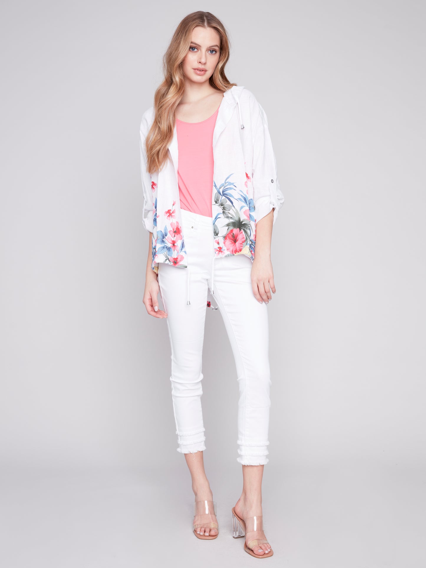 A woman in a white Charlie B floral print linen duster jacket, pink top, and white pants posing against a gray background.