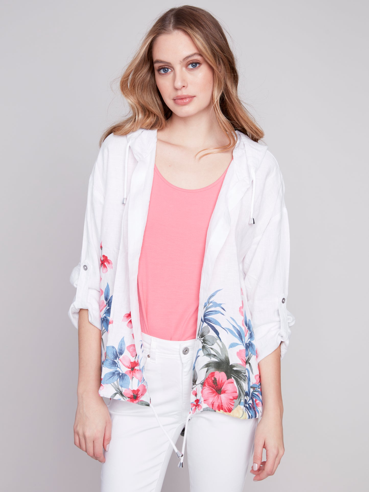 A woman in a white Charlie B floral print linen duster jacket, pink top, and white pants posing against a gray background.