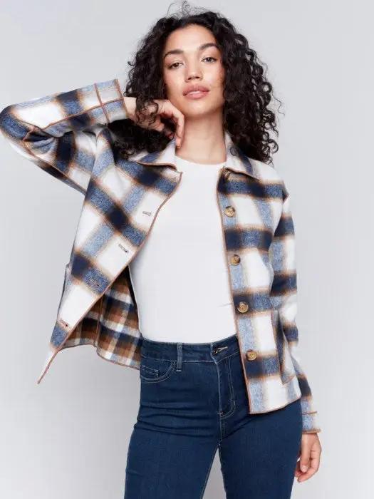 A person with curly hair wearing a white shirt, blue jeans, and a stylish Charlie B Reversible Plaid Jacket poses with one hand touching their neck.