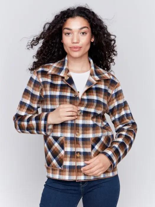 A person with curly hair wearing a white shirt, blue jeans, and a stylish Charlie B Reversible Plaid Jacket poses with one hand touching their neck.