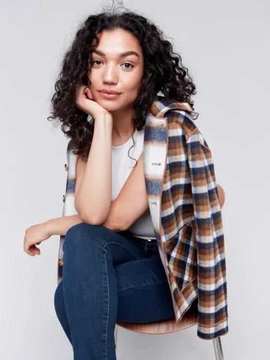 A person with curly hair wearing a white shirt, blue jeans, and a stylish Charlie B Reversible Plaid Jacket poses with one hand touching their neck.