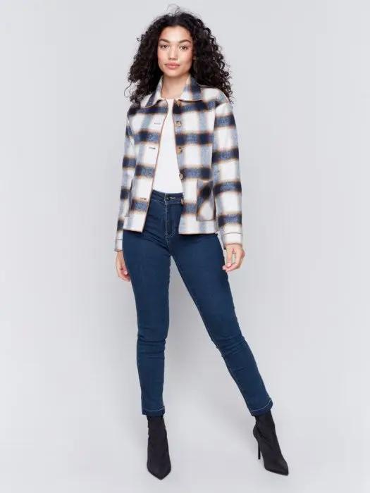 A person with curly hair wearing a white shirt, blue jeans, and a stylish Charlie B Reversible Plaid Jacket poses with one hand touching their neck.