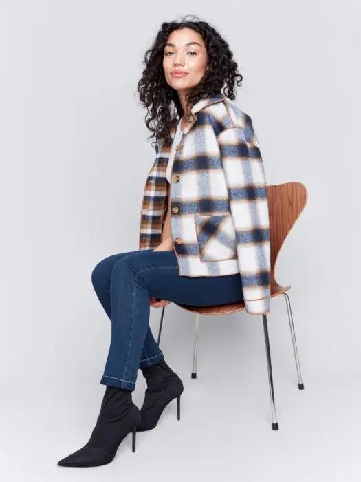 A person with curly hair wearing a white shirt, blue jeans, and a stylish Charlie B Reversible Plaid Jacket poses with one hand touching their neck.