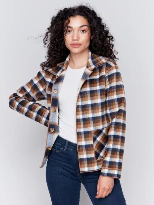 A person with curly hair wearing a white shirt, blue jeans, and a stylish Charlie B Reversible Plaid Jacket poses with one hand touching their neck.