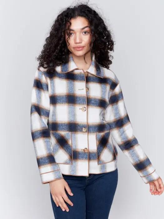 A person with curly hair wearing a white shirt, blue jeans, and a stylish Charlie B Reversible Plaid Jacket poses with one hand touching their neck.