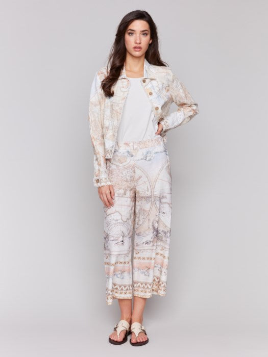 A person stands effortlessly against a plain background, wearing the Charlie B Frayed Linen Jacket and matching pants with a vintage map pattern. The ensemble creates a statement look featuring functional pockets that enhance its unique charm.