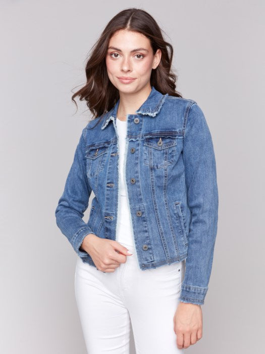 A fashion-forward individual sits on a rattan chair, wearing a chic Charlie B Jean Jacket with Frayed Edges and white pants, their head resting on one hand and a neutral expression.