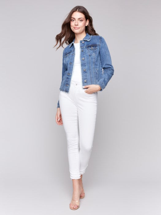 A fashion-forward individual sits on a rattan chair, wearing a chic Charlie B Jean Jacket with Frayed Edges and white pants, their head resting on one hand and a neutral expression.