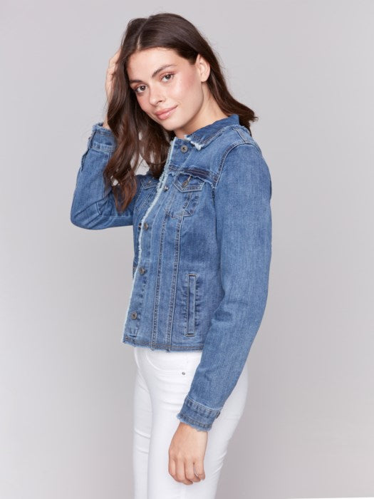 A fashion-forward individual sits on a rattan chair, wearing a chic Charlie B Jean Jacket with Frayed Edges and white pants, their head resting on one hand and a neutral expression.