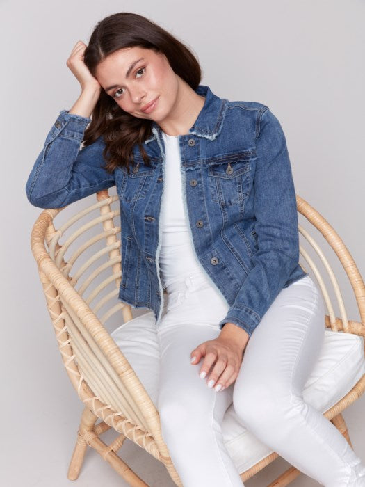 A fashion-forward individual sits on a rattan chair, wearing a chic Charlie B Jean Jacket with Frayed Edges and white pants, their head resting on one hand and a neutral expression.