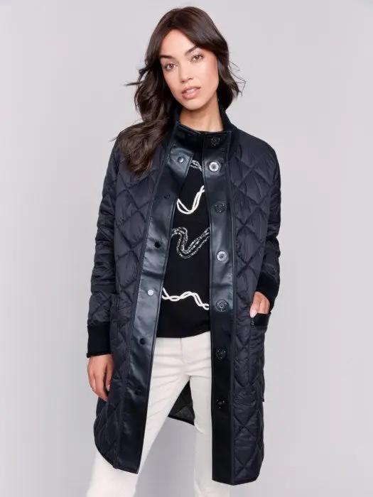 A person with long hair wears a black Charlie B Reversible Quilted Puffer Jacket with a button-up front, a black sweater featuring a white design, and light-colored pants, creating a versatile style perfect for any winter wardrobe. Standing against a plain background, their look is both stylish and adaptable.